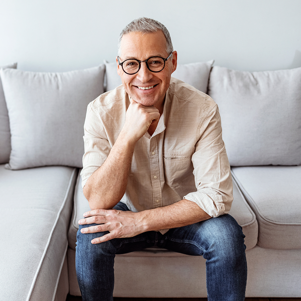 The picture shows a smiling, middle-aged man sitting on a couch, looking directly at the viewer with a relaxed and friendly demeanor.