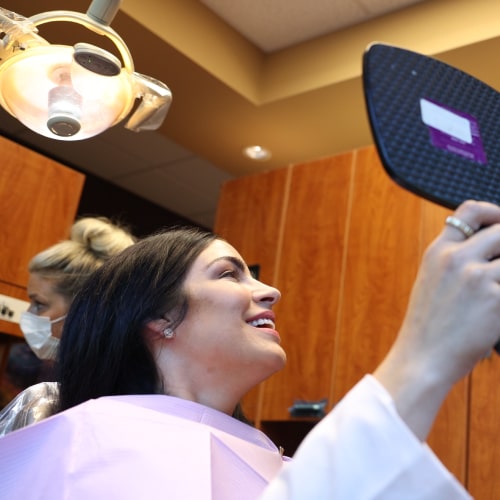 A woman with a small mirror joyfully smiling in a dental chair